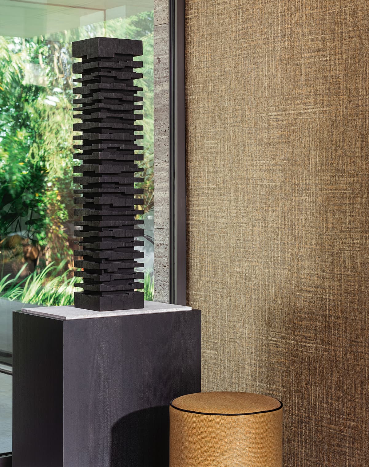 Das Bild zeigt eine schwarze Statue neben einem goldenen Pouf und einer goldenen Tapete. Durch das Fenster fallen viele Grüntöne ins Bild, die für Abwechslung sorgen, während der goldene Pouf besonders heraussticht.