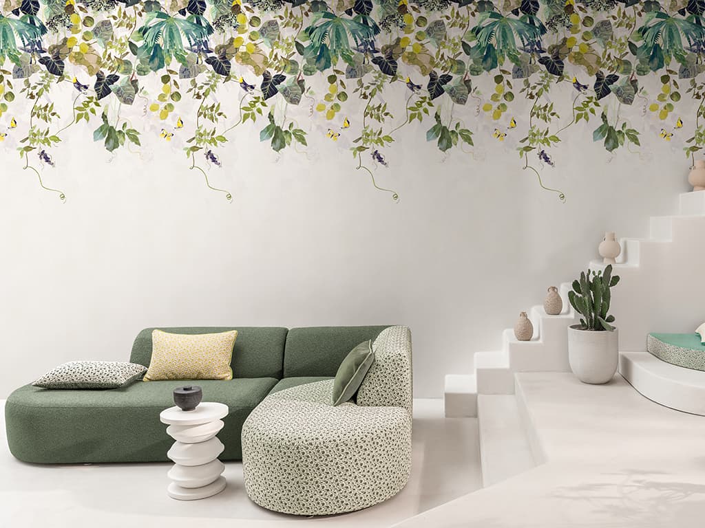 Das Bild zeigt eine Tapete mit Pflanzenprint, während im Vordergrund ein farblich passendes, naturgrünes Sofa steht. Die Farbpalette umfasst viele Grüntöne sowie Gelb und Weiß.