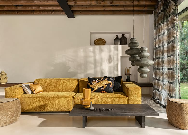 Wohnzimmer mit gelben Sofa, Holztisch, zwei Poufs in und eine gemusterte Gardine