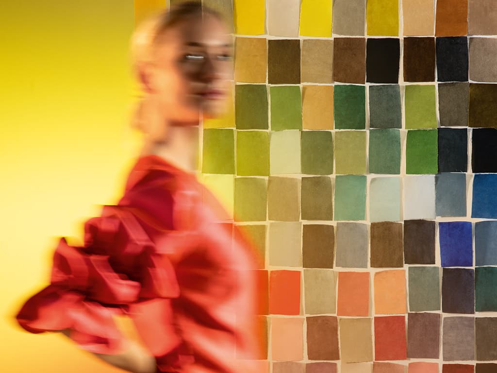 Ein blonde Frau steht vor einer Wand mit bunten Stoffmustern.
