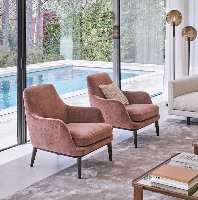 Polstersessel mit rosa Bezugstoff im modernen Wohnzimmer mit großer Fensterfront. Im Hintergrund ist ein Pool.