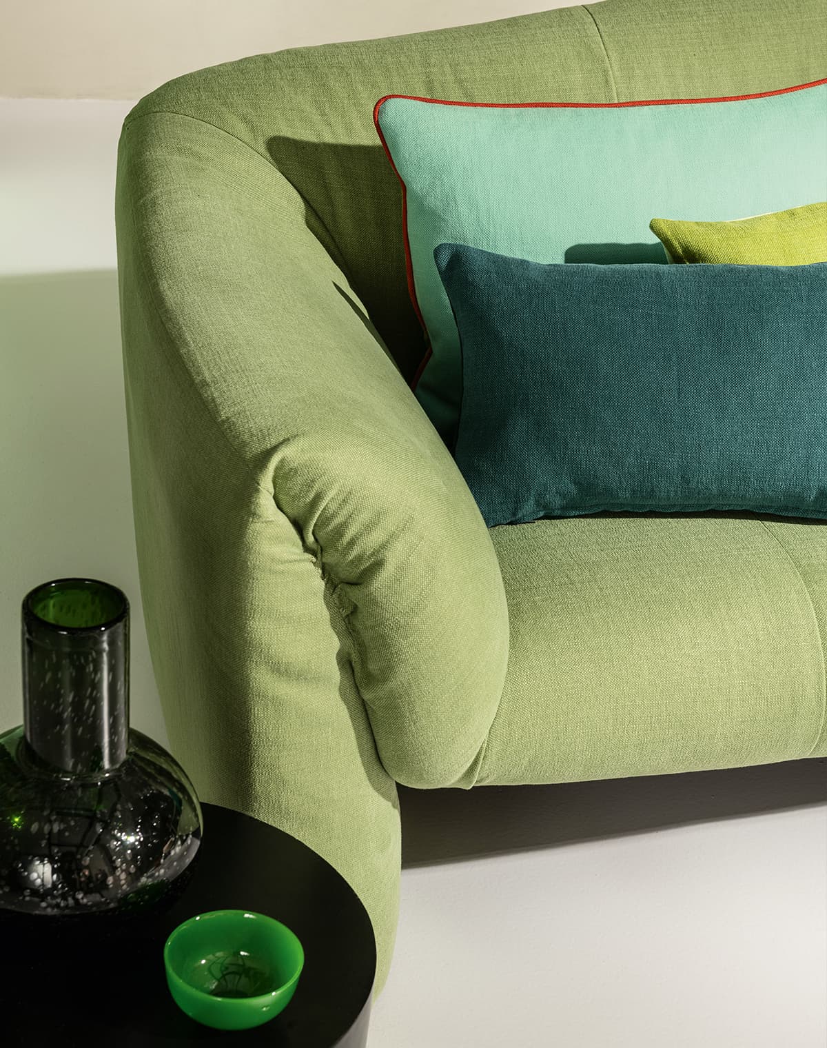 Ein grünes Sofa mit drei Kissen in verschiedenen Grüntönen hinter einem schwarzen Beistelltisch mit einer schwarzen Vase und einer grünen Schale