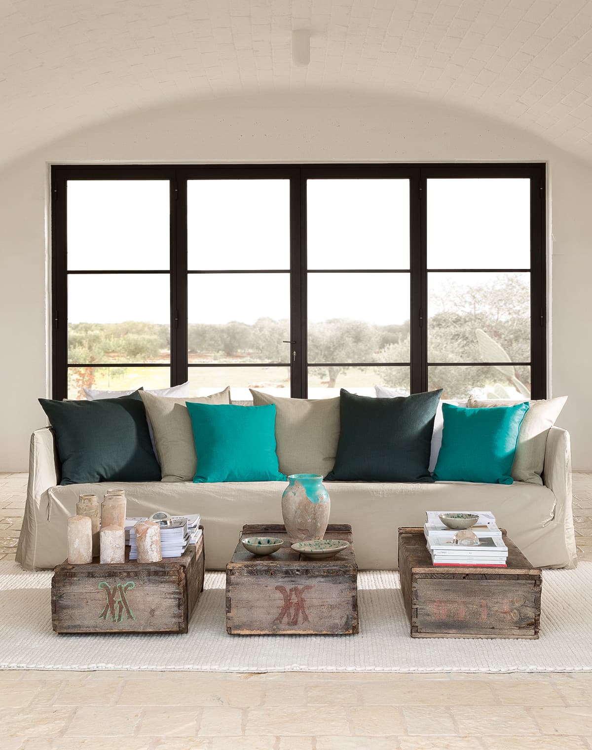 Das Bild zeigt ein Wohnzimmer mit einem weiß-beigen Sofa, einem weißen Teppich und großen Fenstern. Bunte Kissen auf dem Sofa und Altholzkisten als Tisch verleihen dem Raum eine rustikale Note.