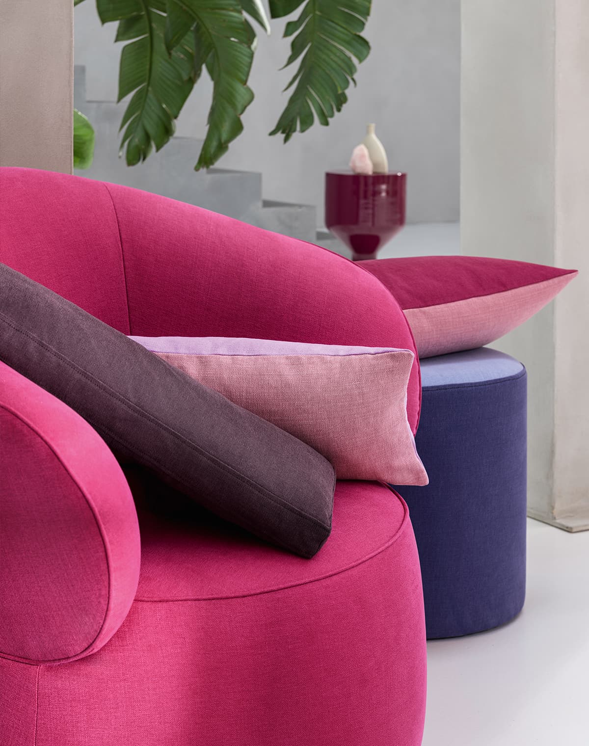 Ein pinker Sessel mit einem braunen und einemrosa Kissen vor einem Lila Pouf mit einem zweifarbigen Kissen in pink und rosa.