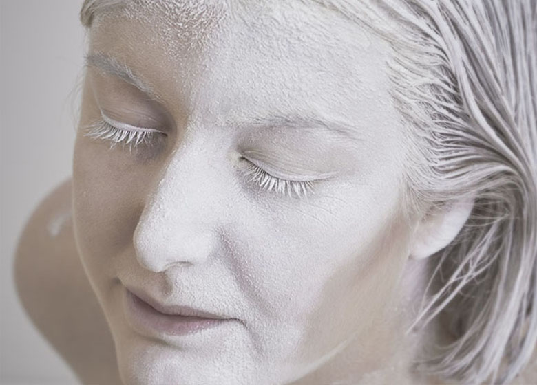 Das Bild zeigt eine Nahaufnahme eines menschlichen Gesichts mit geschlossenen Augen. Das Gesicht ist weiß gepudert, und die sichtbaren Haare sind ebenfalls weiß. Die gesamte Bildkomposition ist überwiegend in Weiß gehalten.