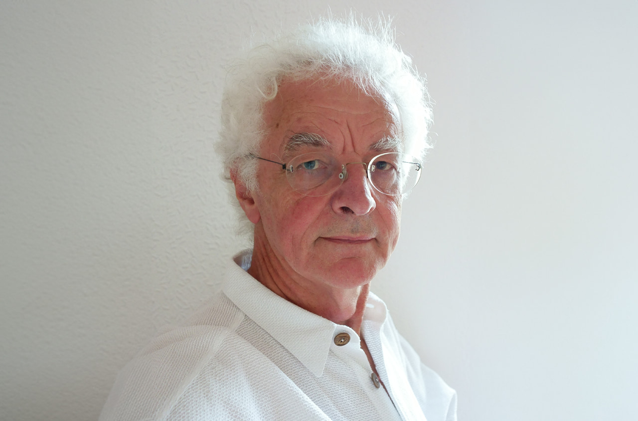 Älterer Mann mit weißen Haaren und Brille schaut in die Kamera- Er trägt ein weißes Hemd.