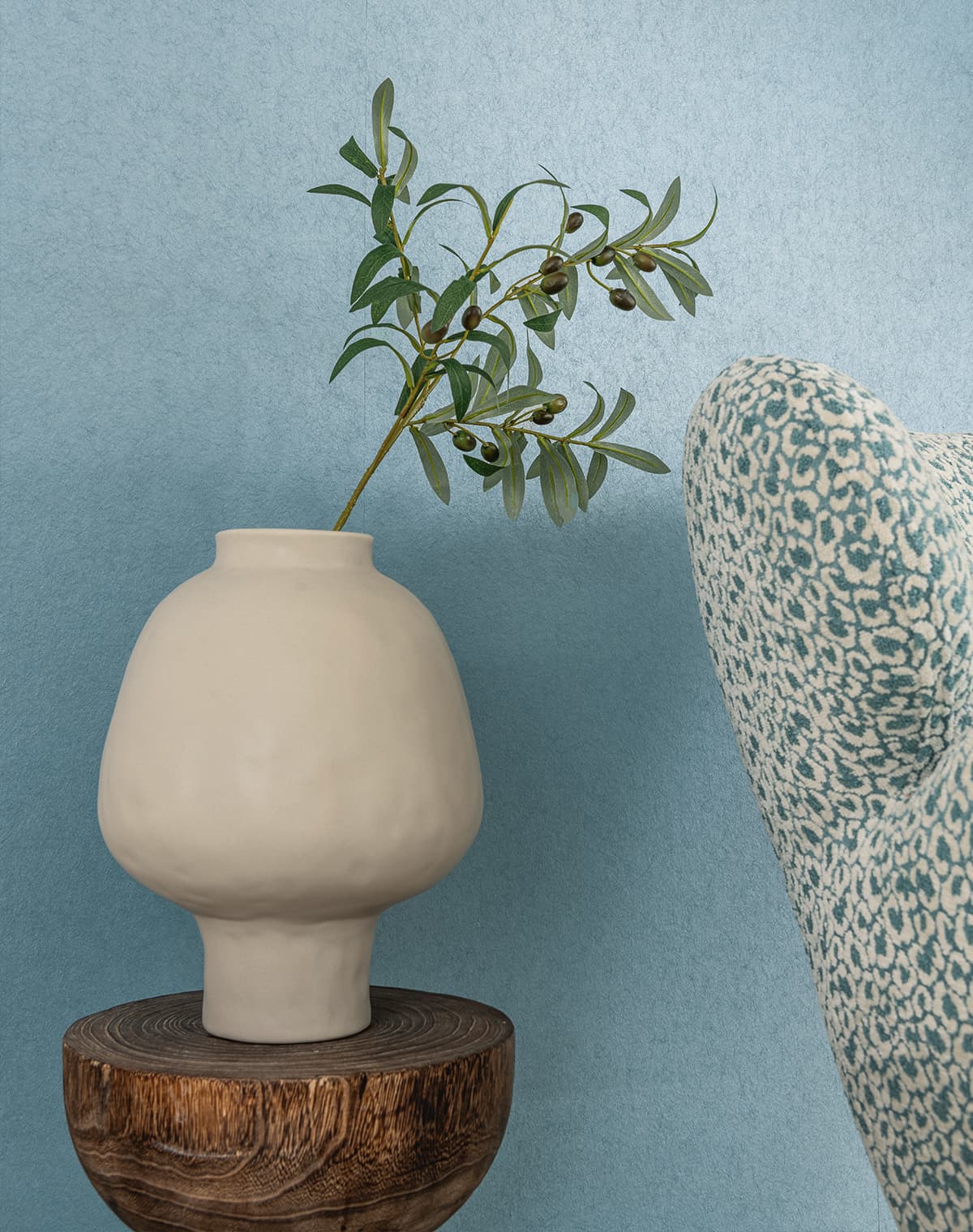Das Bild zeigt ein Wohnzimmer mit blauer Tapete. Ein Naturholztisch trägt eine getöpferten Vase mit einem Olivenzweig. Rechts ist ein Teil eines blau gemusterten Sessels zu sehen, der gut zur Dekoration passt.