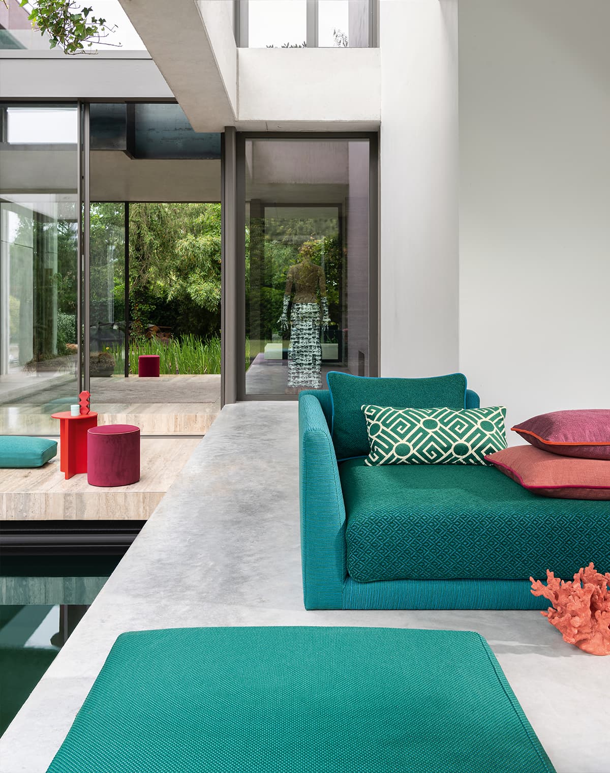 Das Bild zeigt türkis-grüne Outdoor-Möbel am Pool, mit einigen Kissen in Lila und Rot. Die moderne Architektur harmoniert gut mit den Stoffen, deren Farben sich mehrfach im Bild wiederfinden.  