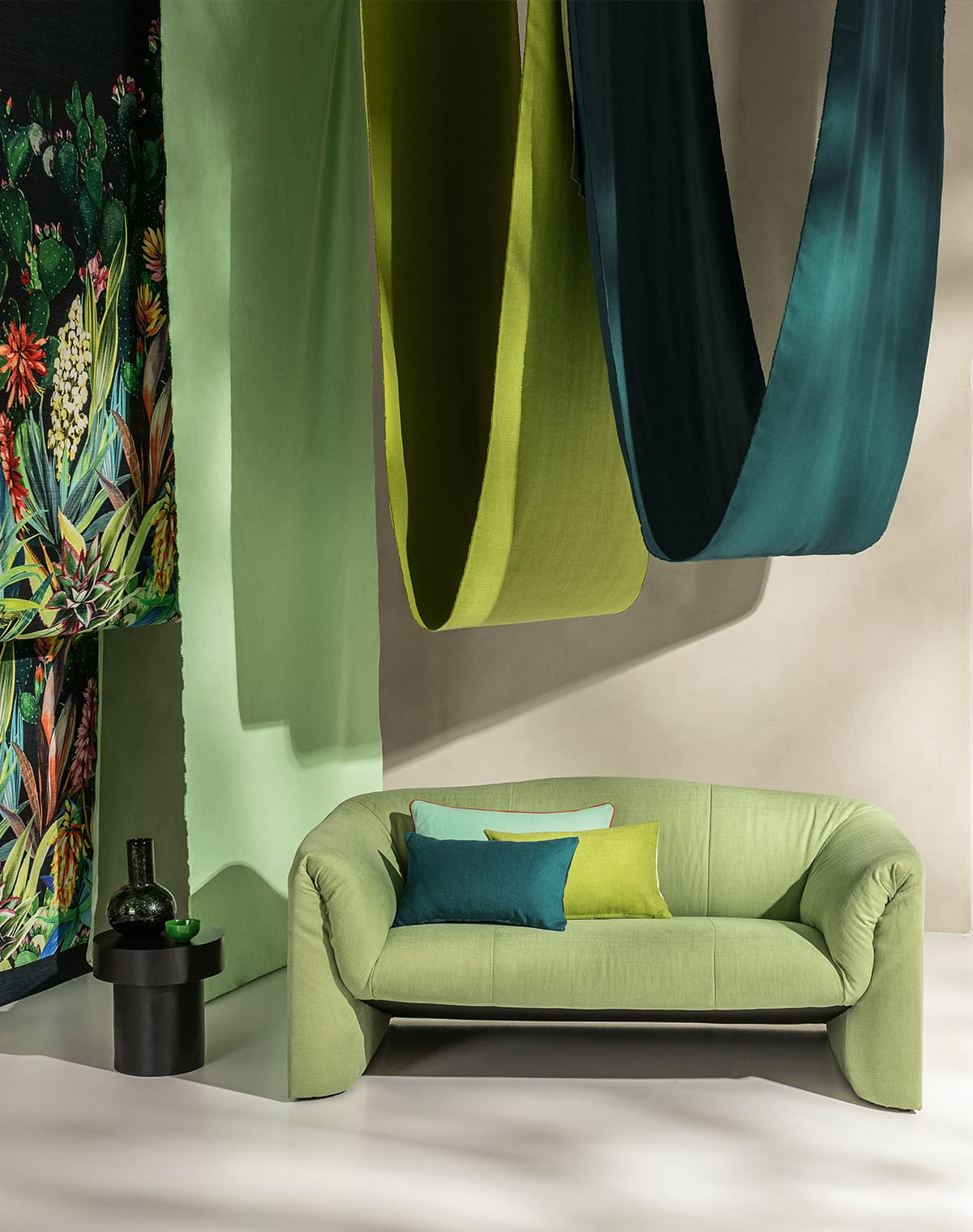 Ein grünes Sofa mit drei grünen Kissen in verschiedenen Tönen. Drei Farbschals von der Decke herabhängend in verschiedenen Grüntönen.Ein Vorhang mit floralem Motiv.