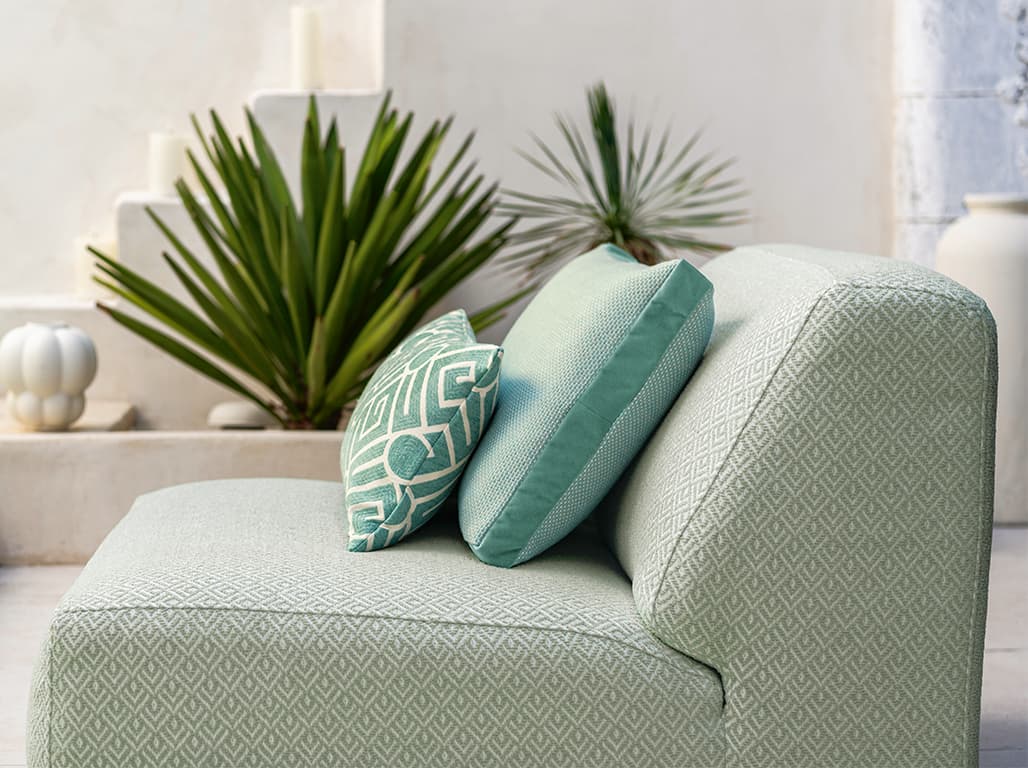 Das Bild zeigt ein seitlich fotografiertes Outdoor-Sofa mit grünen Kissen, die gut zur Bemusterung passen. Im unscharfen Hintergrund sind weiße und grüne Elemente durch Wand und Pflanze zu erkennen, die harmonisch mit dem Sofa wirken.