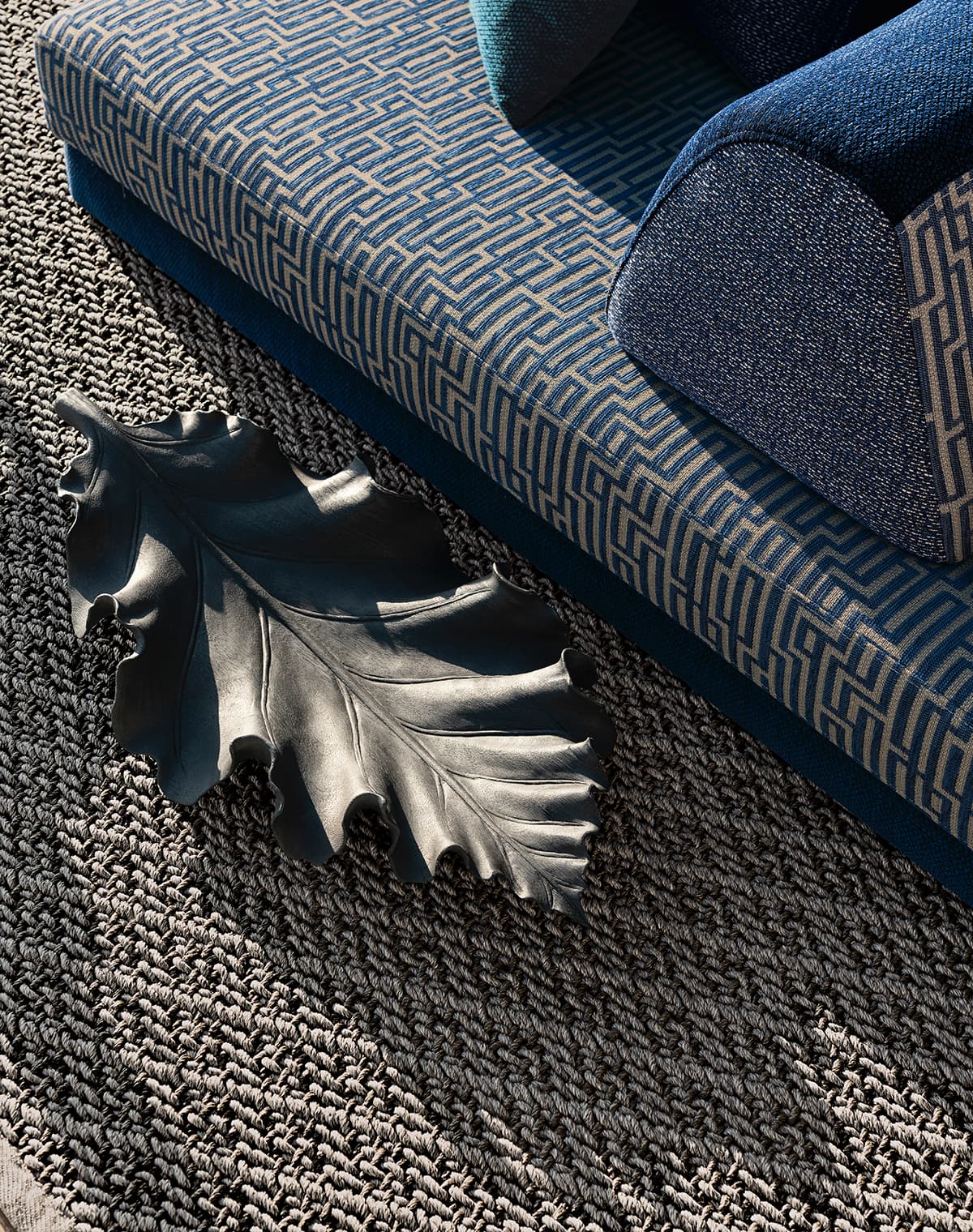Das Bild zeigt eine Detailaufnahme eines leicht braunen Teppichs unter einem blauen Daybed. Darauf liegt ein designvolles, dunkles grünes Blatt, das eine stilvolle Atmosphäre schafft.