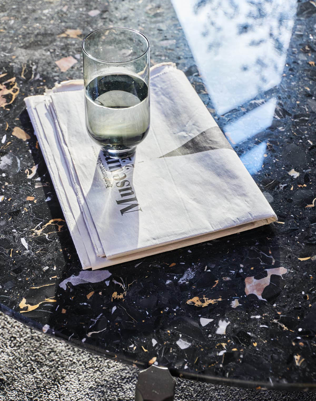Detailaufnahme von dem Tisch Hamilton in Terrazzo Portoro, auf dem eine Zeitung liegt und ein Glas steht