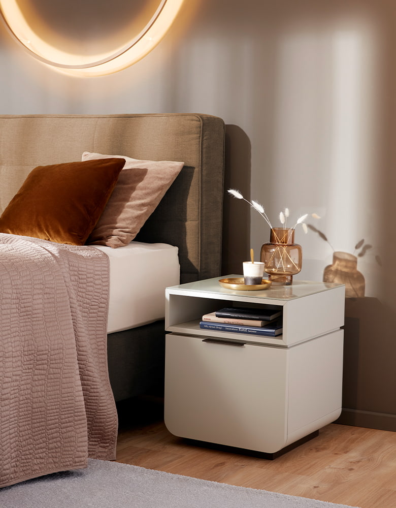 Weißer Nachttisch neben einem Boxspringbett. Über dem Bett hänge eine Ringleuchte.