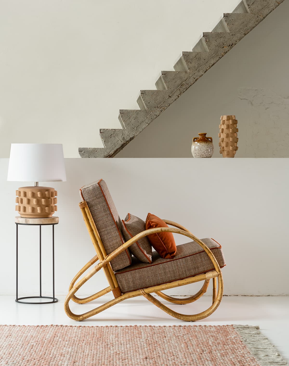Das Bild zeigt im Vordergrund einen Sessel mit Sitzpolstern und Kissen sowie einen orange-weißen Teppich. Im Hintergrund ist eine stilvolle graue Betontreppe zu sehen. Kleine Dekorationsartikel bringen farbliche Abwechslung in den weißen Raum.