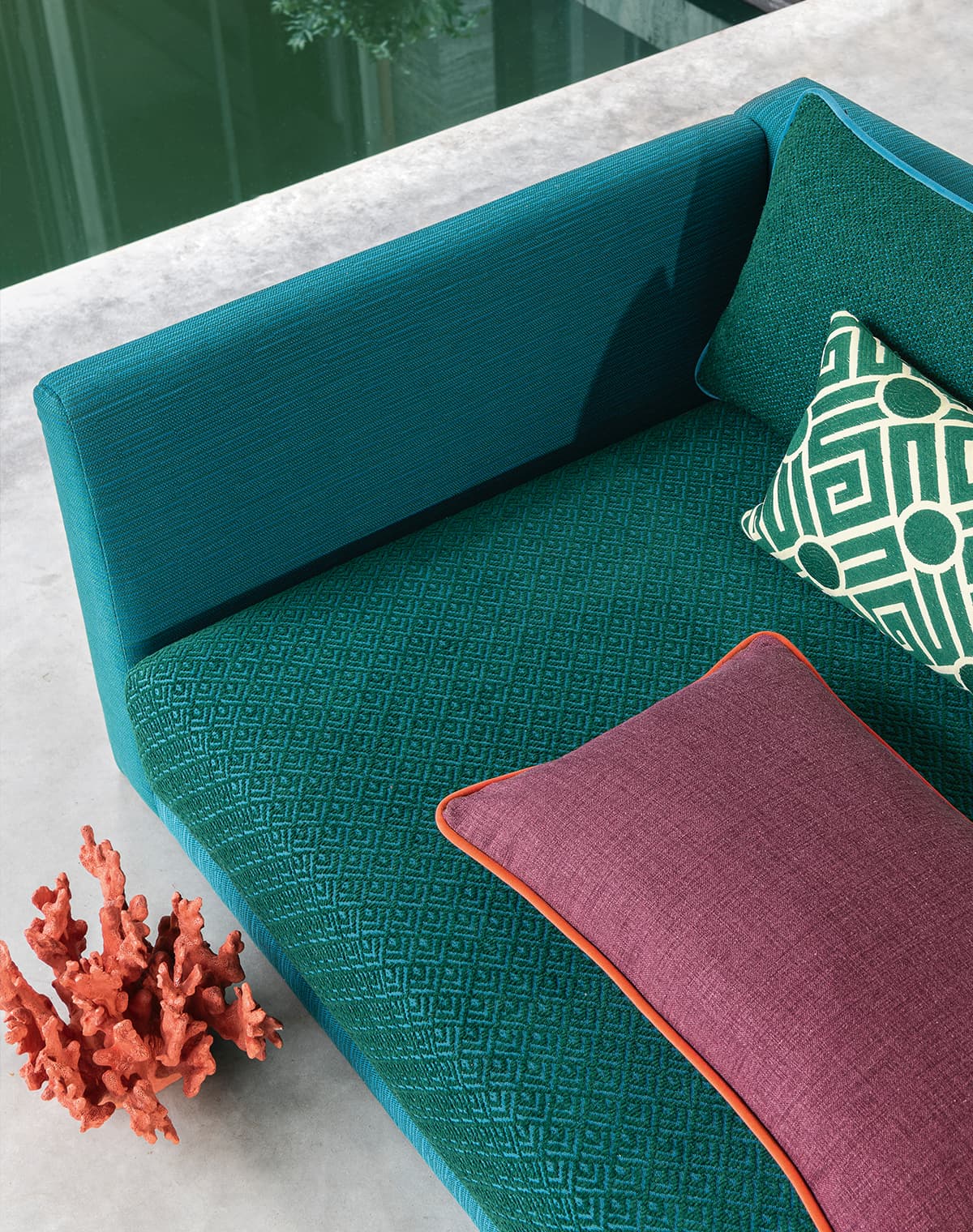 Das Bild zeigt ein grün-türkises Sofa, dekoriert mit rot-lila und gemusterten Kissen. Die Farben der Kissen werden durch ein Dekostück aufgegriffen, während die Grüntöne des Sofas im leicht sichtbaren Naturpool und dessen grünem Wasser reflektiert werden.