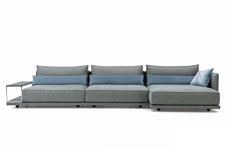 Freisteller eines grauen Sofas mit einer eingebauten Tischplatte und einem blauen Deko-Kissen. An den Rückenlhéhnen liegen blaue Rückenkissen. 