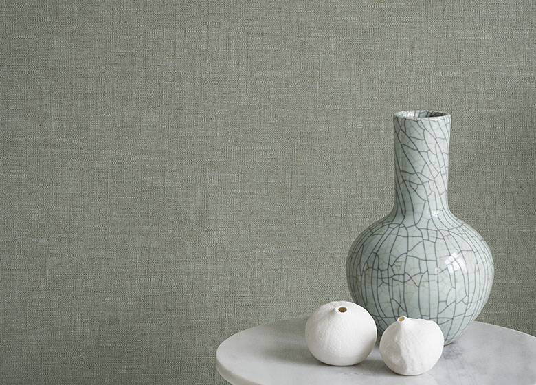 Das Bild zeigt eine Nahaufnahme einer grünen Tapete. Vor der Wand steht ein weißer Beistelltisch mit einer grünen Vase und weißen Dekorationselementen. Das Design und das luftige Grün der Tapete wirken fröhlich und harmonisch.