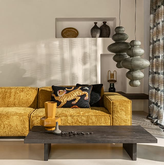 Ein Wohnzimmer mit gelbem Sofa, Holztisch, zwei Poufs in beige und ein gemusterter Vorhang