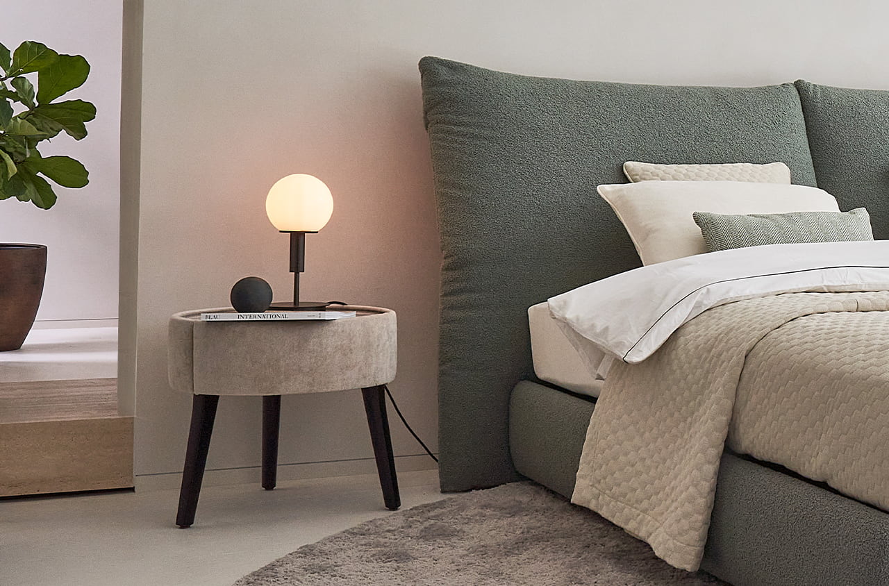Nachttisch Polo in beige, mit einer Lampe drauf, neben einem gürnen Bett