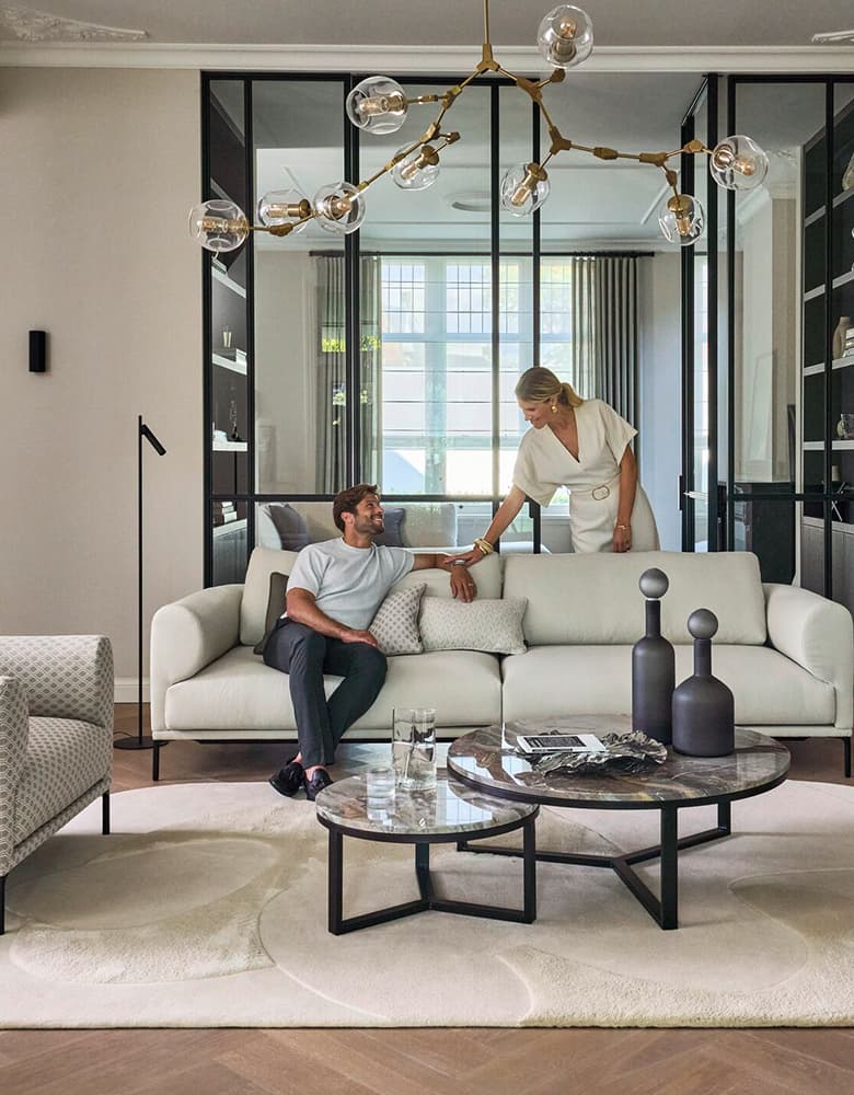 Eine Aufnahme von enem Mann und einer Frau. Der Mann sitzt auf einem weißen Sofa, die Frau steht im, Hintergrund.