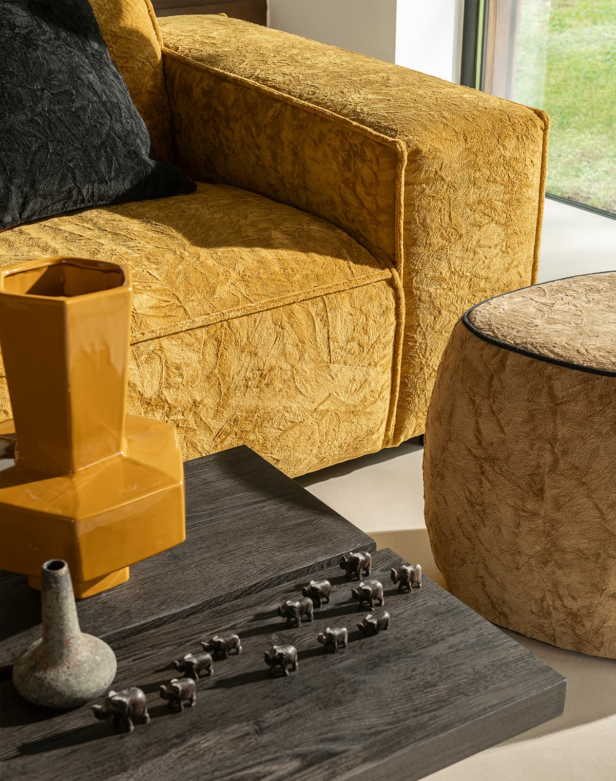 Ein Holztisch, eine gelbe Couch mit schwarzem Kissen und ein brauner Pouf