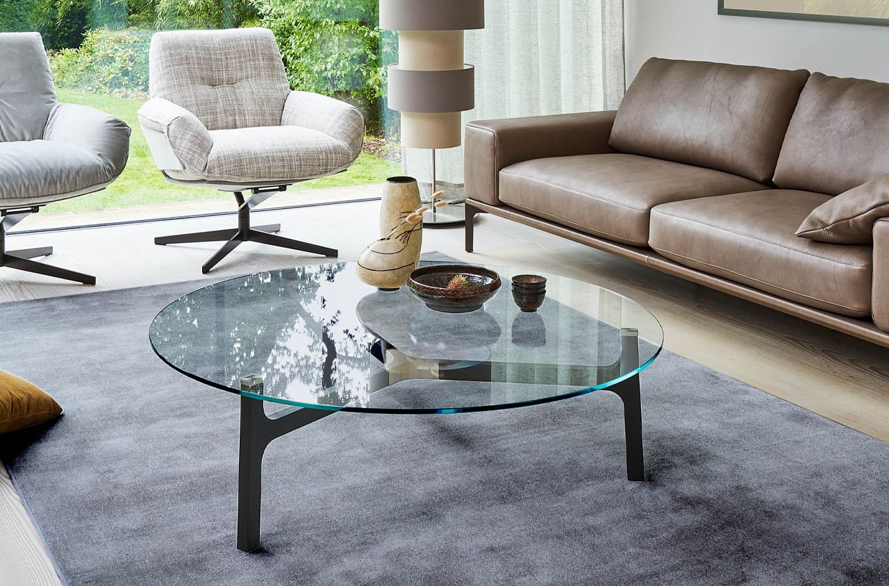 Tisch Porto mit Glasplatte in einer Wohnsituation auf einem Teppich und mit zwei Sesseln und einem Sofa dahinter