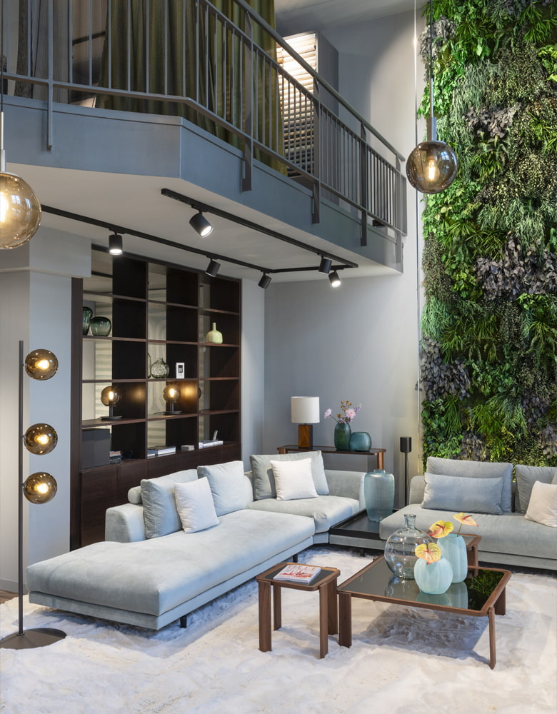 Großes Sofa im Showroom, dabei stehen zwei Couchtische und eine Stehleuchte. Im Hintergrund ist eine Pflanzenwand zu sehen. 