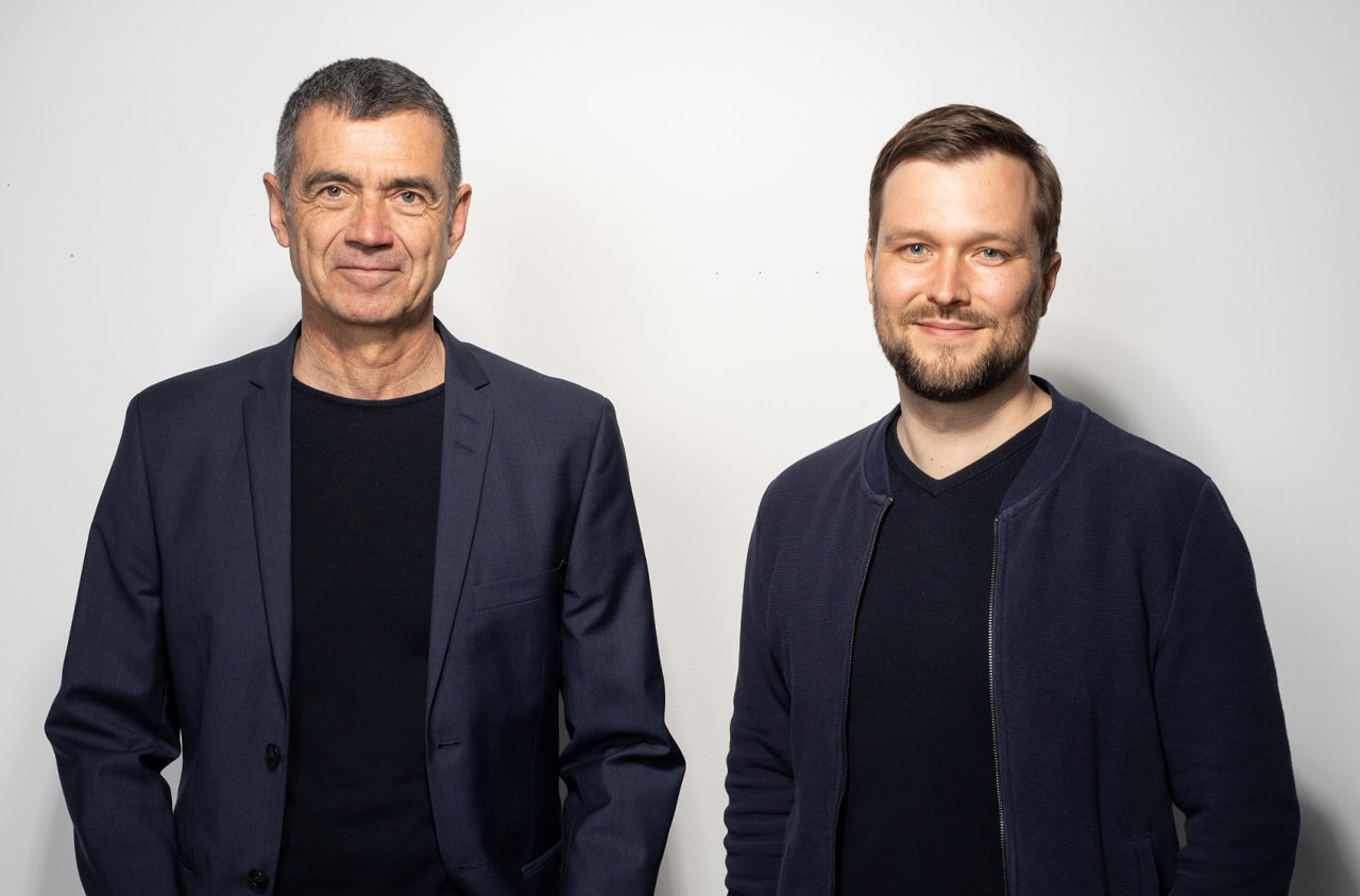 Portraitfoto von zwei Männern die freundlich in die Kamera schauen. Beide tragen dunkle Kleidung.