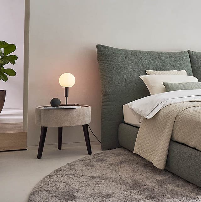 Nachttisch Polo in beige mit schwarzen Füßen, auf dem eine Leuchte steht und welcher neben einem Bett steht