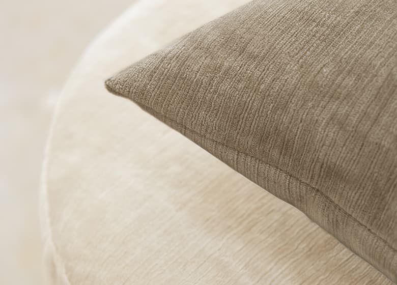 Das Bild zeigt eine Detailaufnahme eines beigen Kissens. Die gestreifte Struktur des Stoffes ist deutlich sichtbar und harmoniert gut mit dem darunterstehenden Poufhocker.