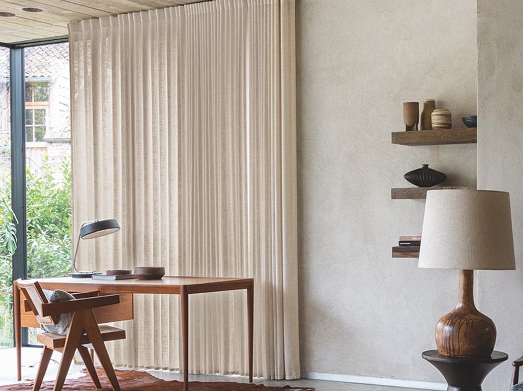 Das Bild zeigt einen Arbeitsplatz vor einem beige-weißen Vorhang. Der Schreibtisch aus dunklem Holz bildet einen Kontrast zur beige-weißen Wand im Hintergrund. Der Raum wirkt ruhig und harmonisch.