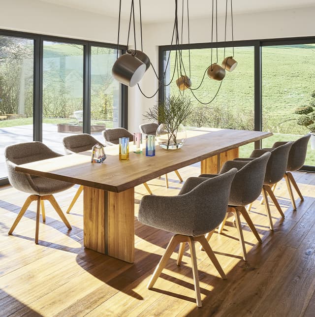 Braune Stühle um einen Esstisch aus hellem gelblichen Holz, im Hintergrund sind große Fenster