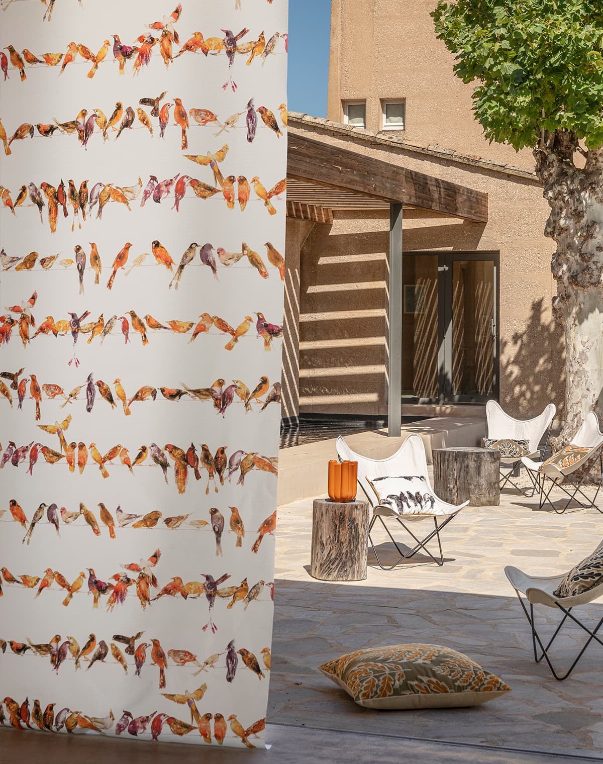 Ein weißer Vorhang mit Vogel-Print und weiße Stühle im Outdoorbereich mit gemusterten Kissen.