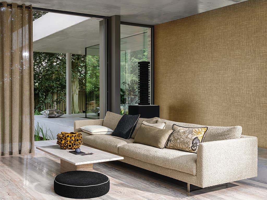 Das Bild zeigt ein großzügiges Sofa vor einer goldenen Tapete in Beige- und Brauntönen. Auf dem Sofa liegen Kissen in verschiedenen Farben und Mustern, während der glänzende Steinboden und die Architektur dem Raum Eleganz verleihen.