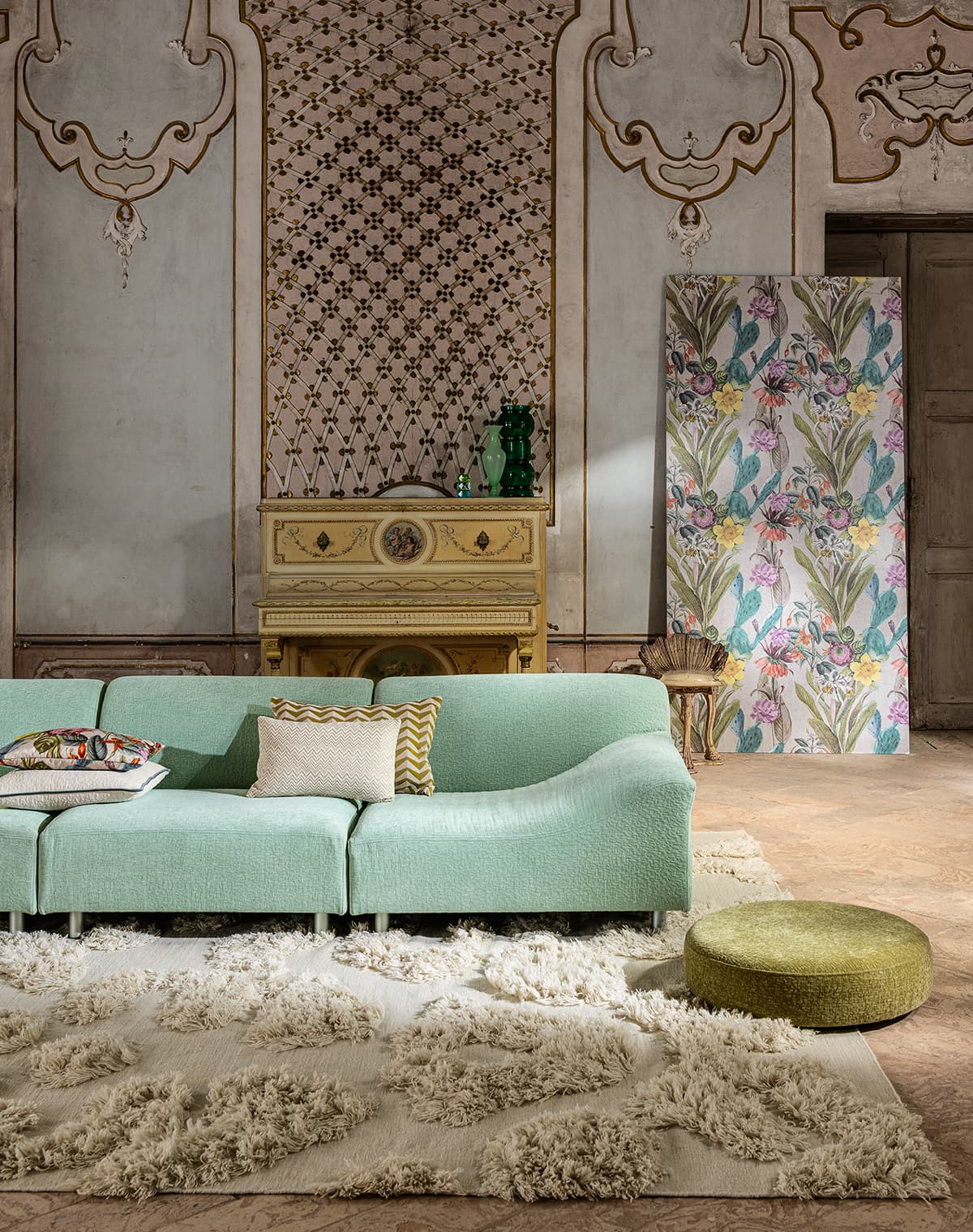 Ein grünes Sofa auf dem 4 Kissen liegen, ein weißer Teppich, ein grüner Pouf und eine Tapete mit floralem Muster